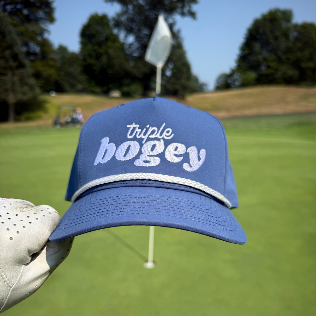 Blue cap with 'triple bogey' text held by a gloved hand on a golf course.